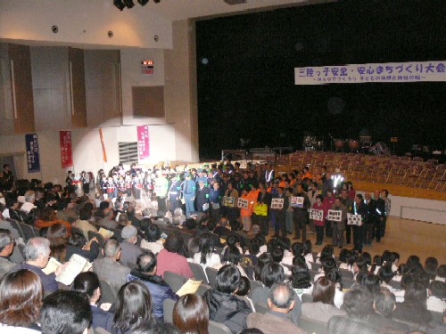 多数の参加者が集まったホールの大きな舞台の前で、関係者が整列している様子を観客席の後方から撮影した写真