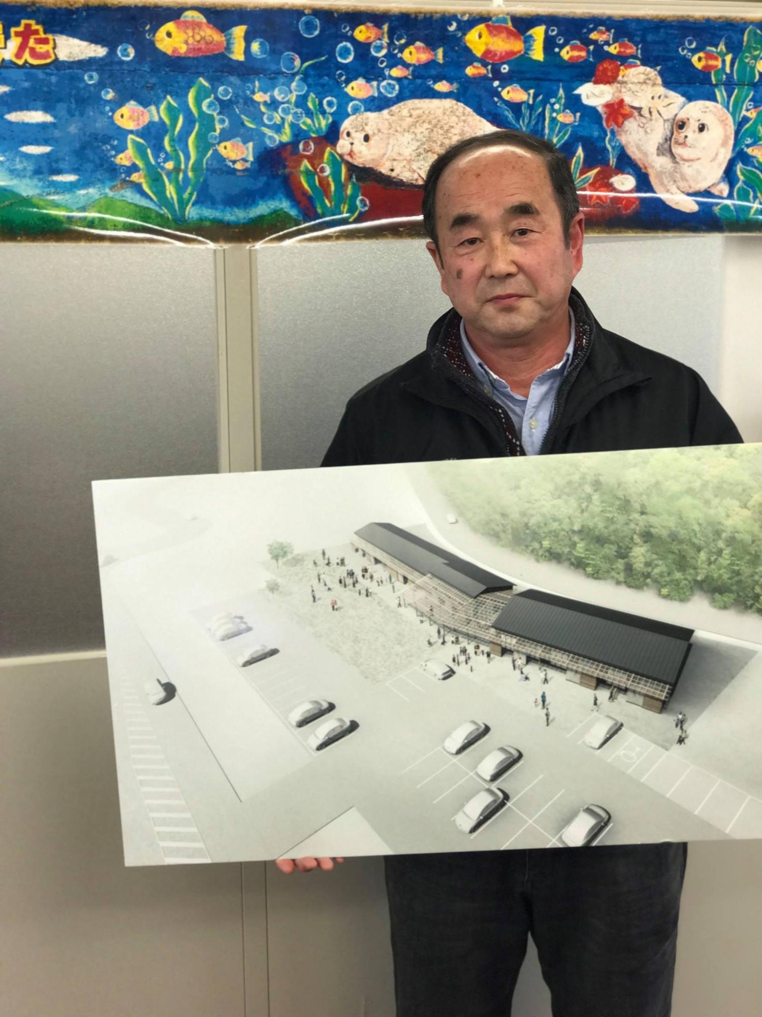魚やアザラシ、海藻が描かれたポスターが貼られたガラス戸の前に、広い駐車場がある横長の商業施設のイメージ図のパネルを持った男性が立っている写真
