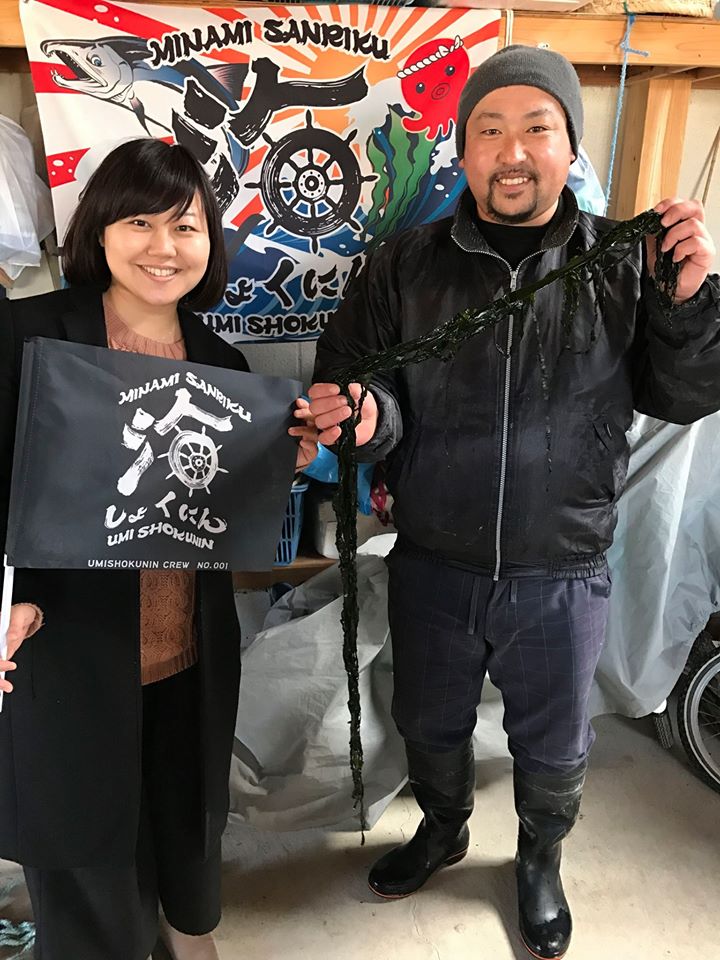 大漁旗を背景に「海しょくにん」と書かれた黒い旗を持った女性リポーターと、長い生のわかめを手に持った男性が並んで立っている写真