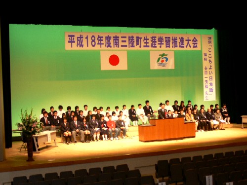 「平成18年度南三陸町生涯学習推進大会」と書かれた横断幕が掲げられたホールのステージ上で、スーツ姿の関係者の男性が演台で話をし、制服姿学生や参加者が両隣に設置された椅子に座って話を聞いている様子の写真
