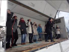 ステージの上に10人ほどの男女が上がり、前に出た男性と後ろに並んだ女性の一人がマイクを持って歌っている様子の写真