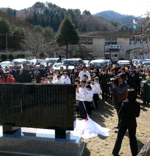 黒い大きな記念碑の前で、大勢の子どもたちを含む関係者が集まり、報道関係者の前で式典が行われている様子の写真