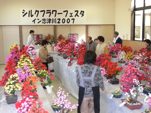 「シルクフラワーフェスタイン志津川2007」の看板が掲げられた屋内の会場で、来場した多くの人々が、赤、ピンク、黄色など鮮やかな色合いの造花で作られたアレンジメント作品を、興味深げに鑑賞している様子の写真