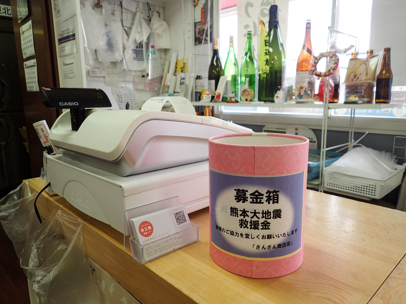 商店のカウンターのレジ横に設置されたピンク色の熊本地震救援金募金箱を写した写真
