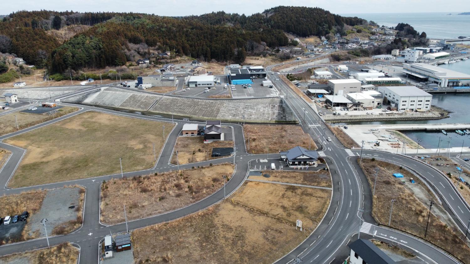 海に向かって走る道路沿いに建設された防潮堤と漁業施設が見え、周囲に整備された区画と低木が点在する志津川湾岸地域の昼間の風景を高台から定点カメラで撮影した写真