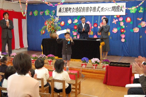 青い幕で装飾されたステージに、「南三陸町立港保育園卒園式並びに閉園式」と書かれた横断幕が掲げられ、関係者などが拍手をする中、一人の園児が賞状を持って壇上に立っている式典の様子の写真