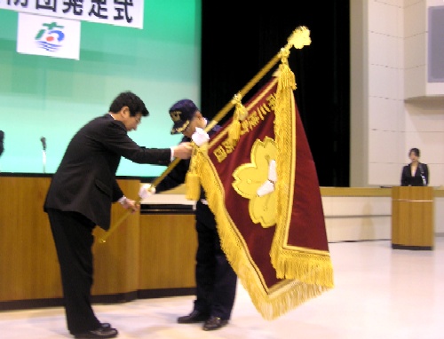 会場のステージ上で、町長が、消防の制服を着た男性に旗を手渡している発足式の様子の写真