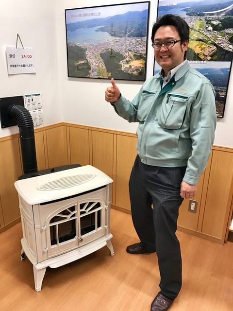 航空写真などが壁に飾れれ、白い薪ストーブが置かれた部屋で、右手の親指をあげたポーズをして立っている佐藤克哉さんの写真