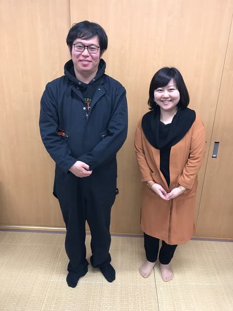 木目調の扉の前に笑顔で並んで立っている女性リポーターと佐藤太一さんの全身写真