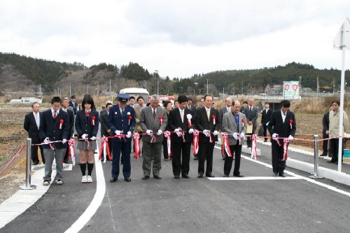 多数の来賓や関係者が、白いテープに赤いリボンをあしらった開通式のテープカットラインを横一列に並んで持っている式典の様子の写真
