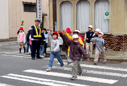 小学生たちが数名で横断歩道を渡る練習をしており、子供たちは右手を高く上げて横断の意思を示し、交差点の安全を確保するために誘導している制服姿の警察官が奥に立っている交通安全教室の様子の写真