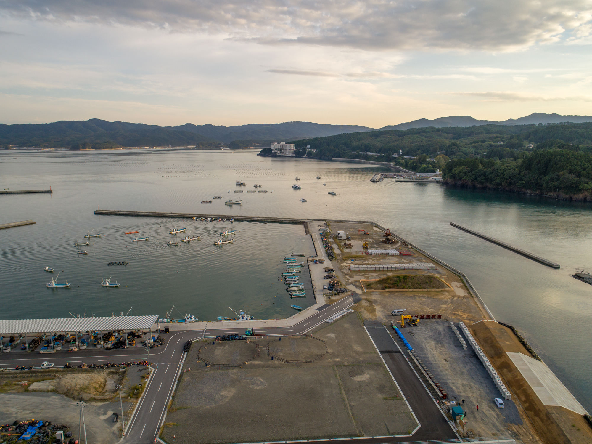 手前に防波堤と整備中の岸壁が見え、その奥には漁船が多数停泊し、右奥には入り江と山林が広がる港の写真