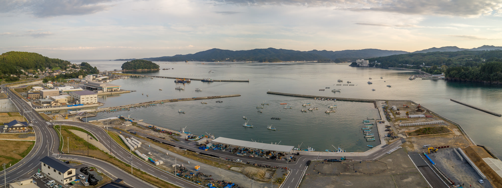 港全体を上空から広く見渡し、左に山と住宅地、中央に港と防波堤、右奥に山々と湾が広がる様子を夕方に撮影した港の写真