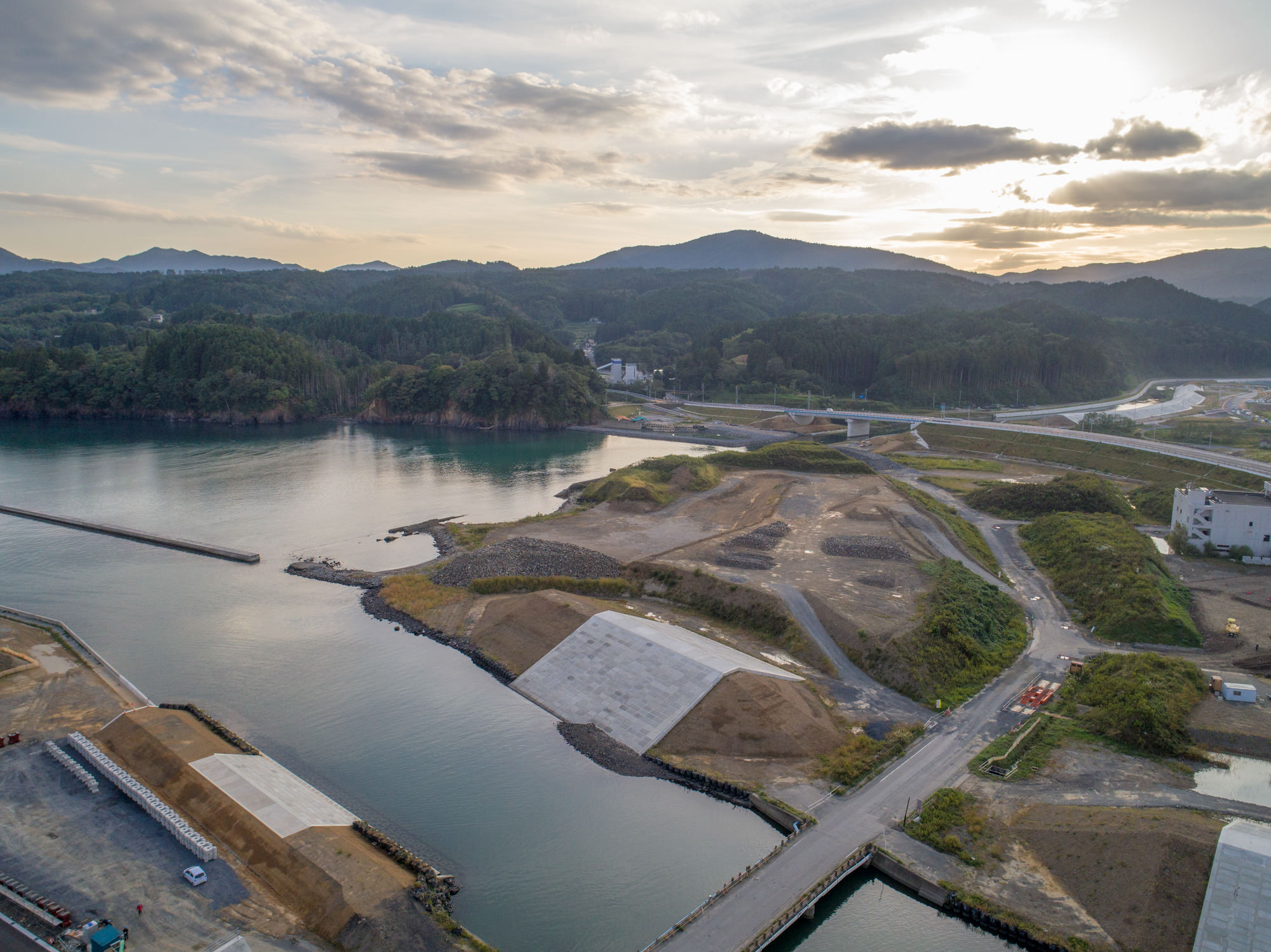 手前に湾と防潮堤があり、奥に広がる山林と造成中の土地、その奥にかかる橋と夕日が重なる気仙沼市唐桑町の防潮堤付近の写真