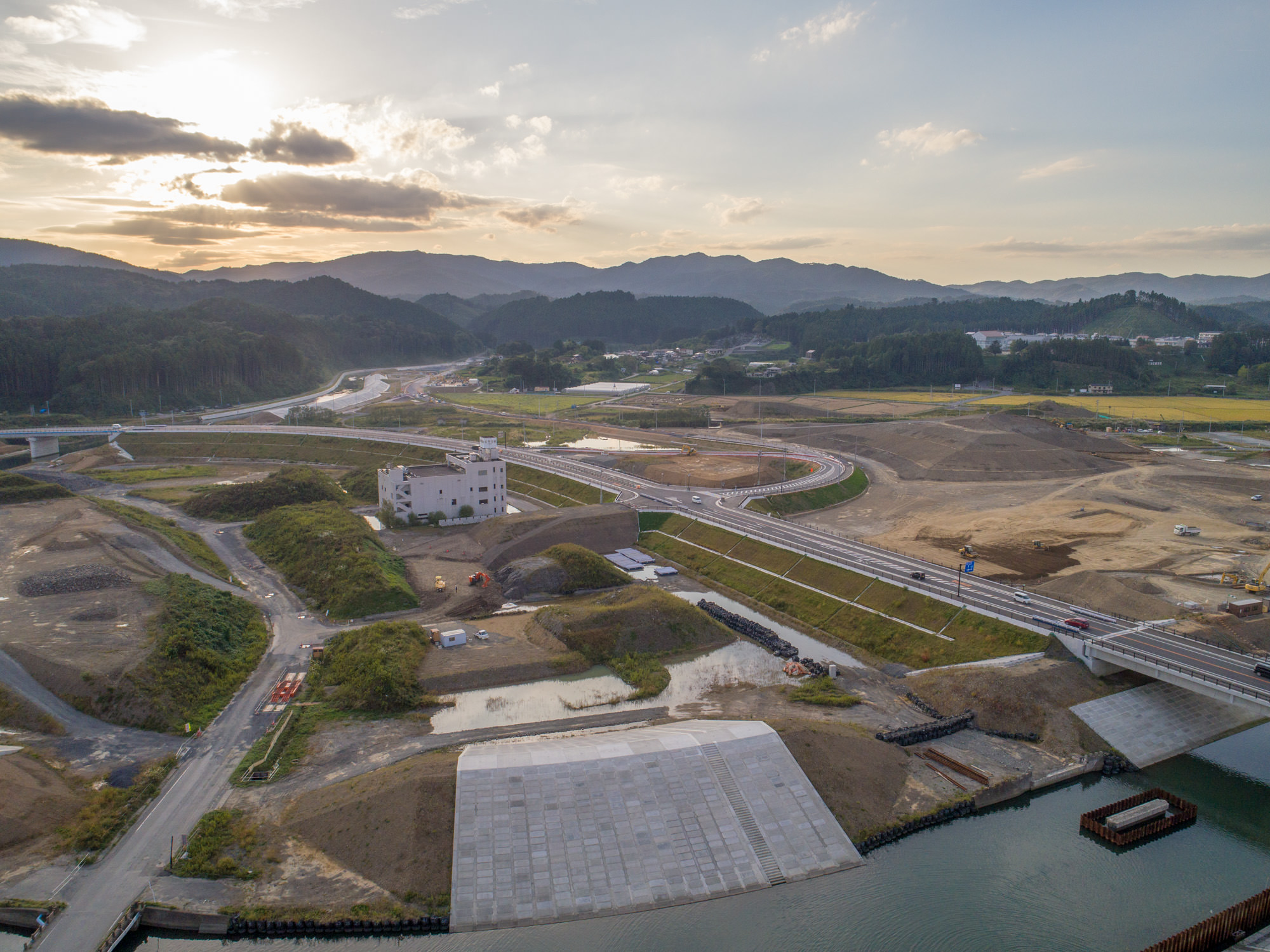 堤防の手前から奥に向かって盛土と整備中の土地が広がり、中央に白い建物、その先に道路や車が行き交う様子が見える復興造成地の写真