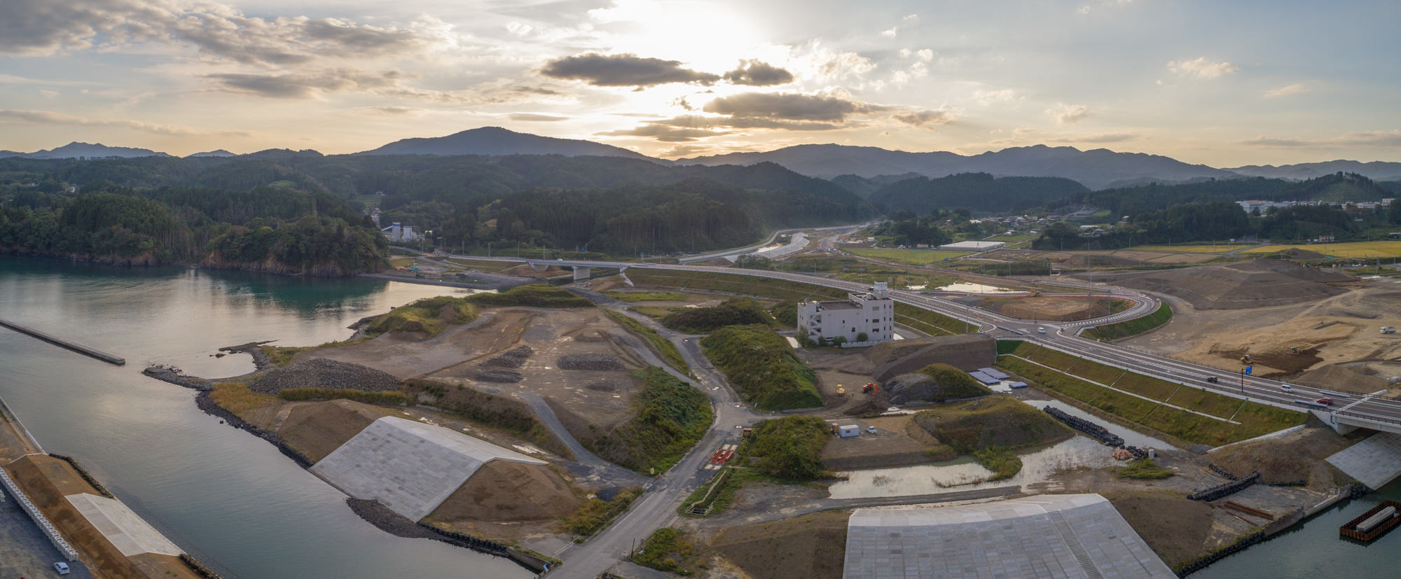 湾の右側から内陸に向かって道路と造成中の土地が広がり、夕日を背にした山並みが背景に広がっている防潮堤付近の写真