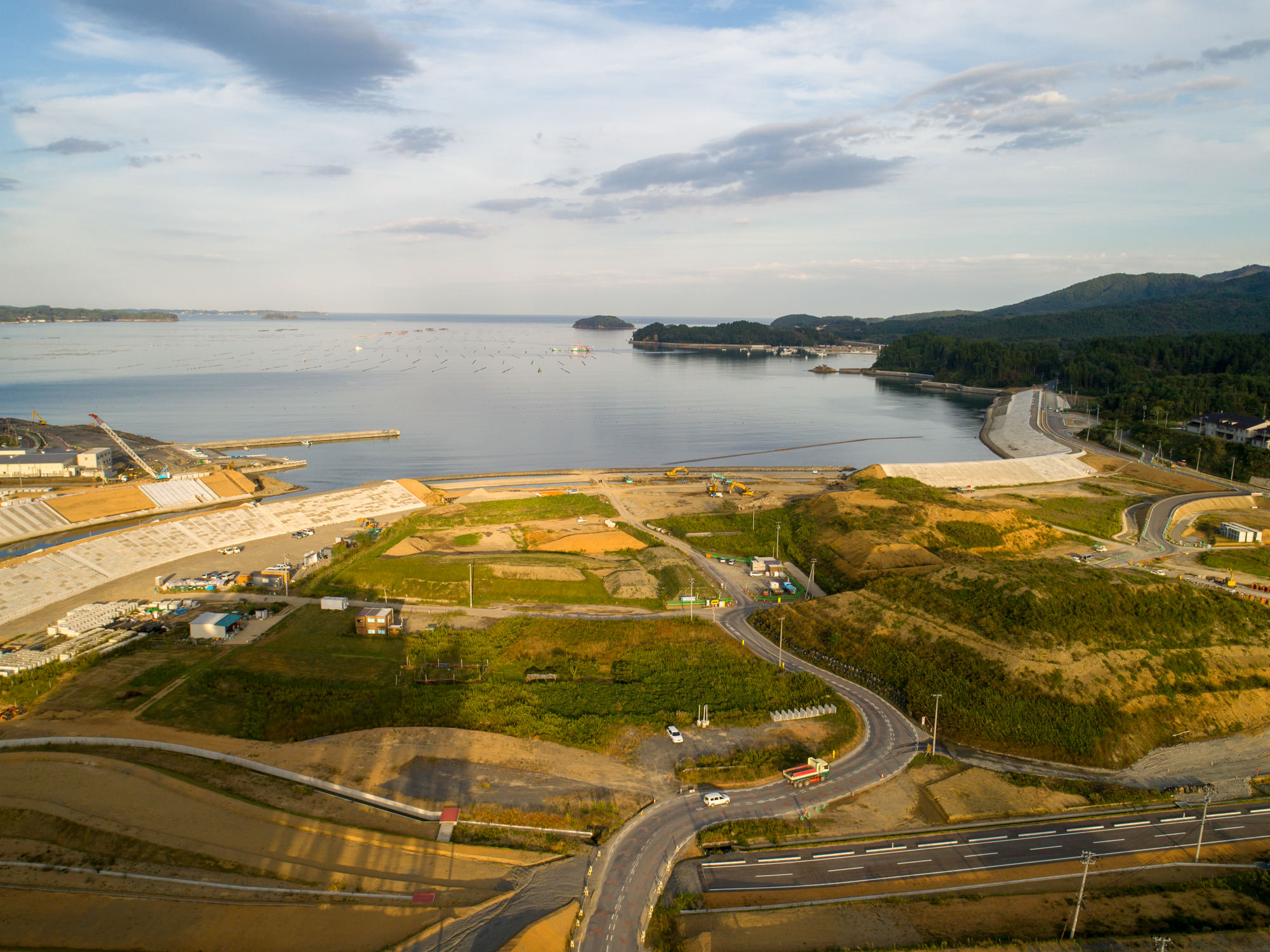 建設中の橋の手前を車道が横切り、工事中の河岸と周囲山地が見える近景全体の写真