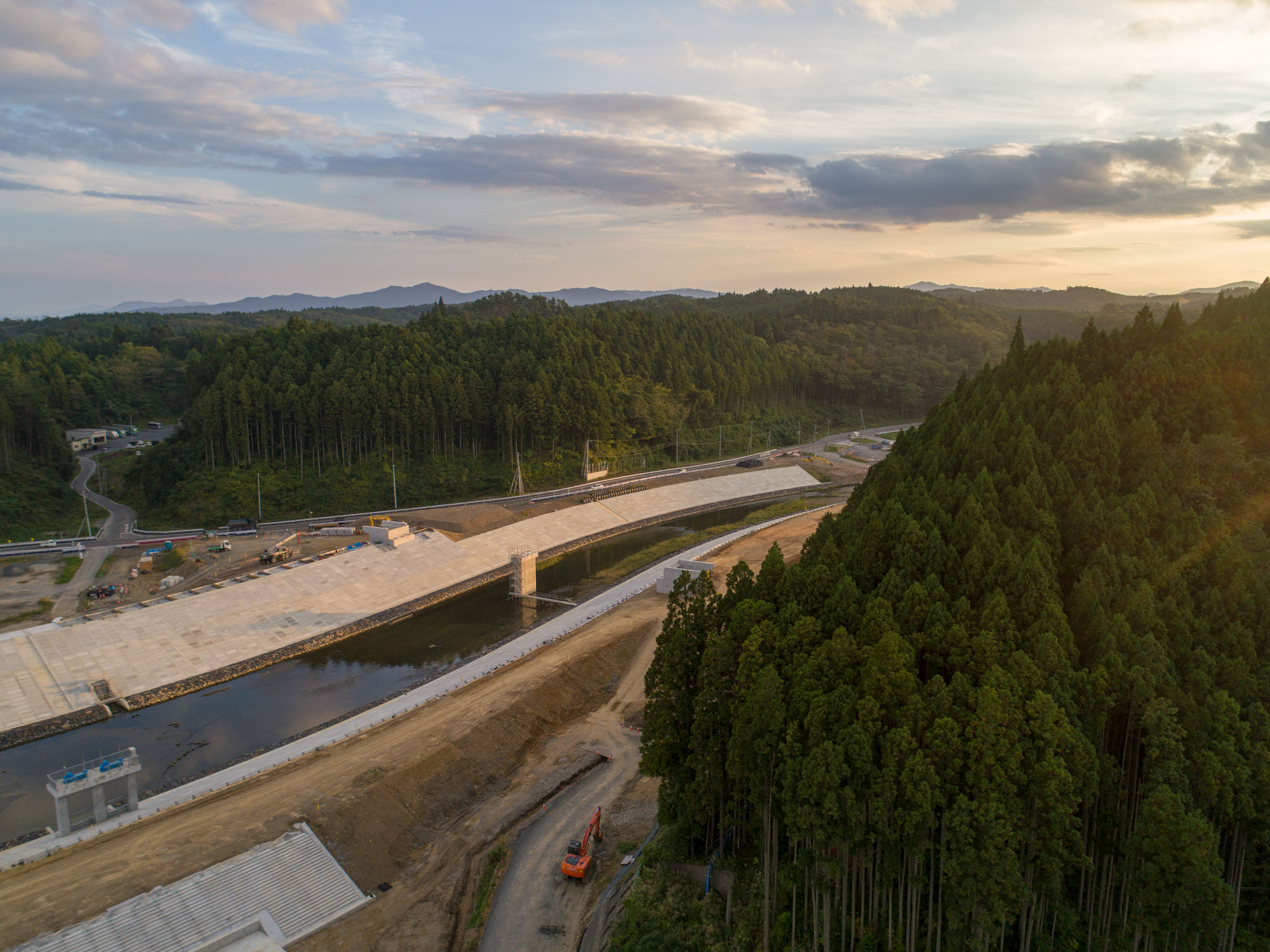 右手前の山林と奥に広がる山々を背景に、川沿いに建設中の堤防と道路が続く様子を高所から見下ろした写真