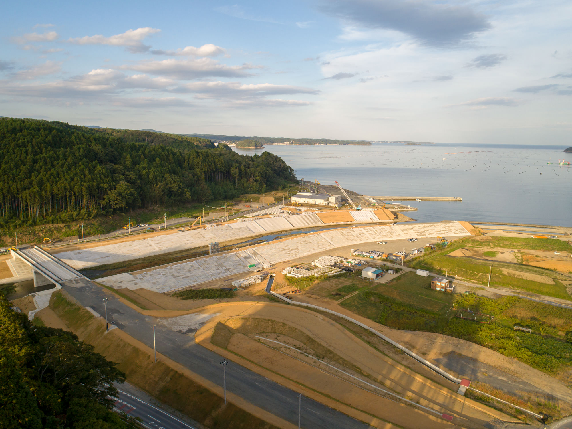 埋立地造成中の河岸と斜面法面、重機が並ぶ工事現場と湾を含む景観の写真