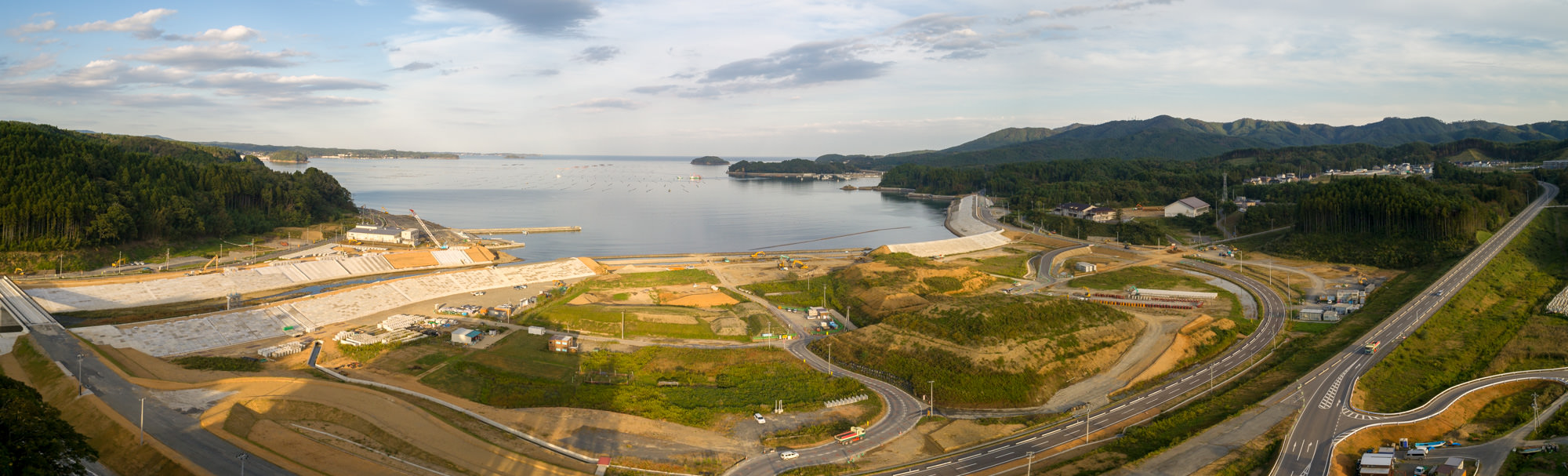 海岸近くの広い埋立地と造成地、湾奥に海と小島、林と山並みが見えるパノラマ全景の写真