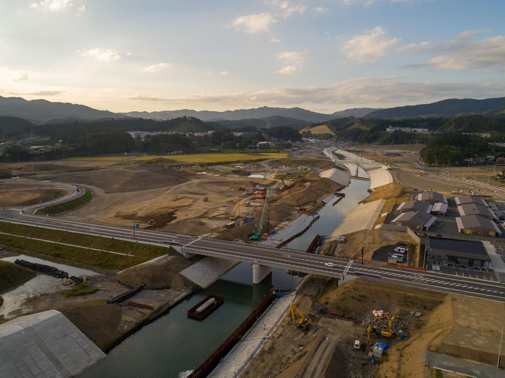 橋の近くで工事中の河岸、奥に店舗風建屋と道路・駐車場が写り込んだ全景の写真