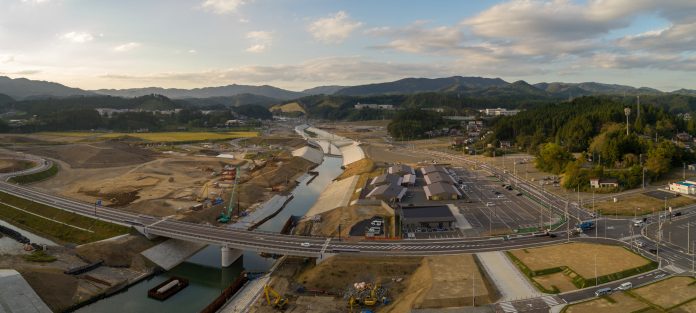 建設中の橋と川、広い舗装済み道路と駐車場、周囲に山並みが広がる全景の写真