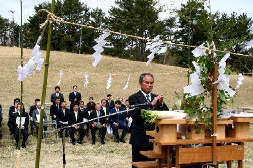 冬枯れの芝生の広々とした屋外で、神事が行われており、スーツ姿の男性が祭壇の前で柏手を打ち、後方に多くの関係者が着席している様子の写真