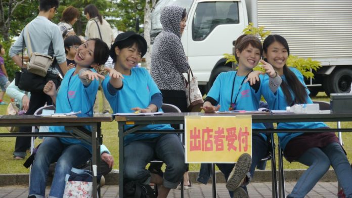 屋外イベントの出展者受付で青いTシャツを着た4人のスタッフが笑顔で座り、手を前に出してポーズをとっている写真
