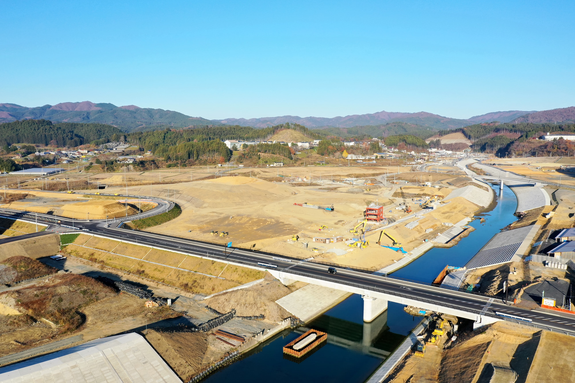 広々とした工事現場の中を川がゆったりと流れ、その川の上には橋が架けられていて、周囲には開発が進む様子が見える写真