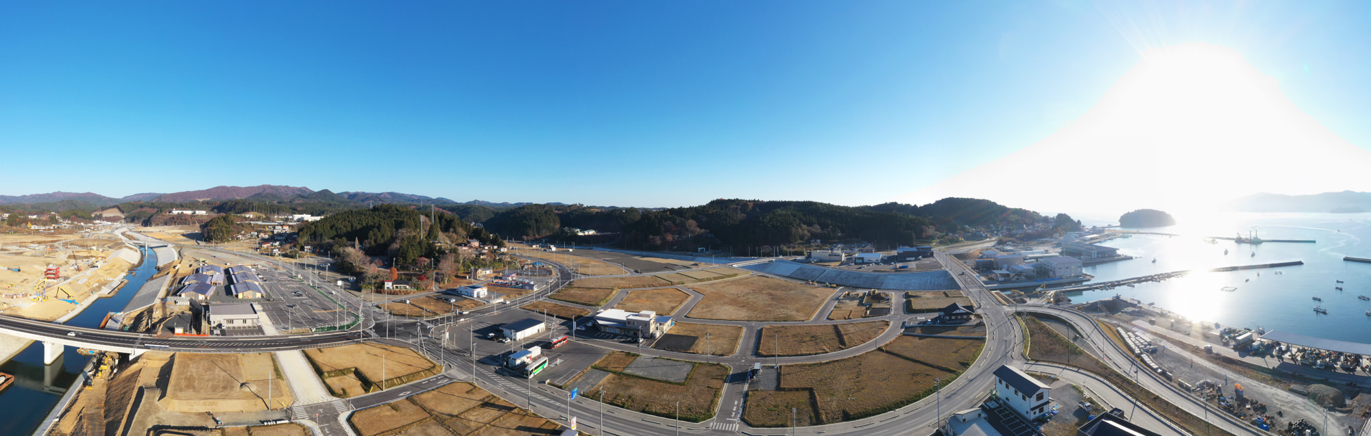青空の下、穏やかな海と緑に覆われた山々に囲まれた港町が広がり、建物や道路が整然と配置されている様子が一望できる写真