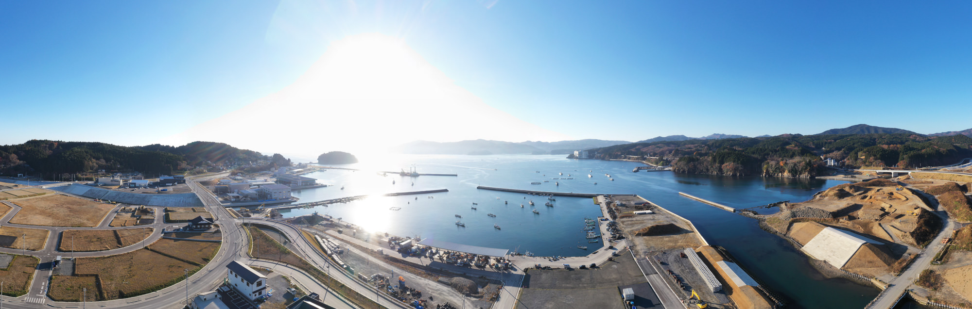 青空の下、穏やかな海に面した港湾地域が広がり、道路や建物が整然と並び、複数の船舶が停泊している様子が見える写真