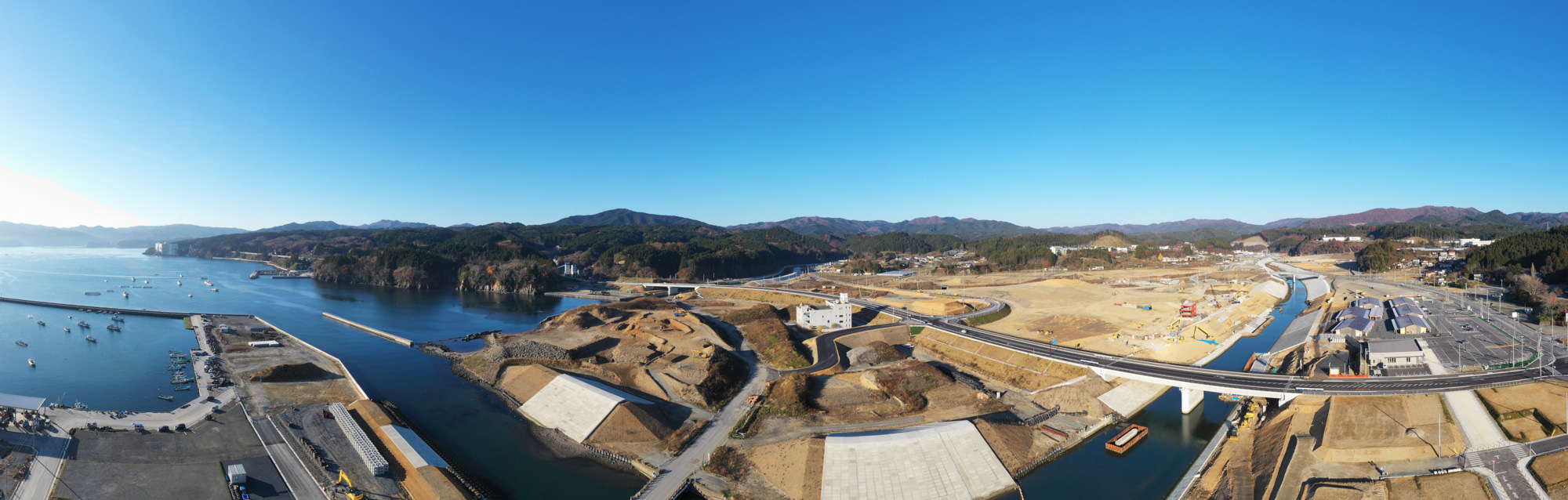 青空の下、海と山に囲まれた広大な土地に橋や道路が建設されており、自然の中でインフラ整備が進められている様子が見える写真