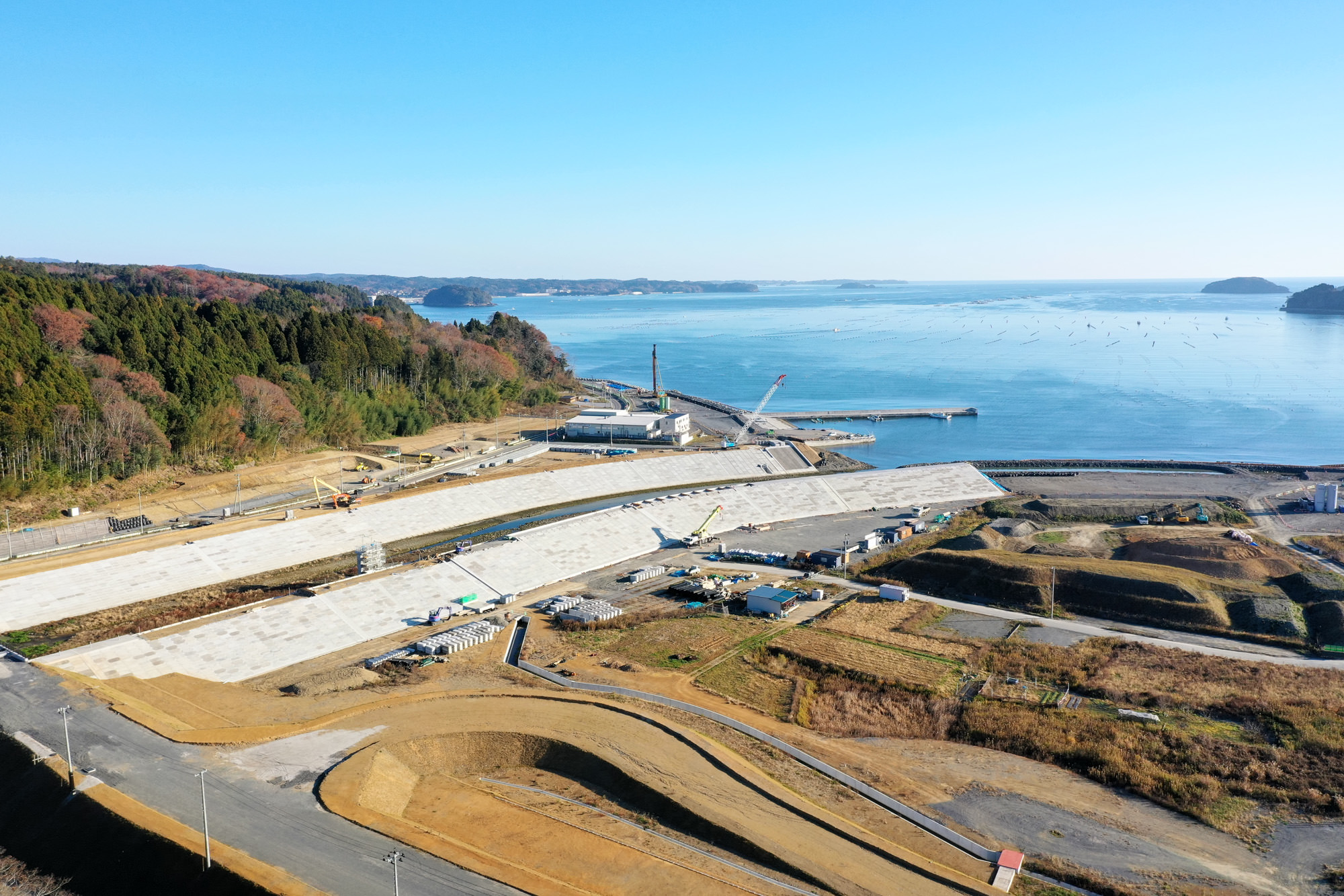 海沿いの開発中の風景として、大型のコンクリート構造物やクレーンが並ぶ建設現場があり、片側には森林が広がり、もう片側には海が広がっていて、複数の工事車両や仮設の建物が点在し、平地と緩やかな丘が混在する地形が見える写真