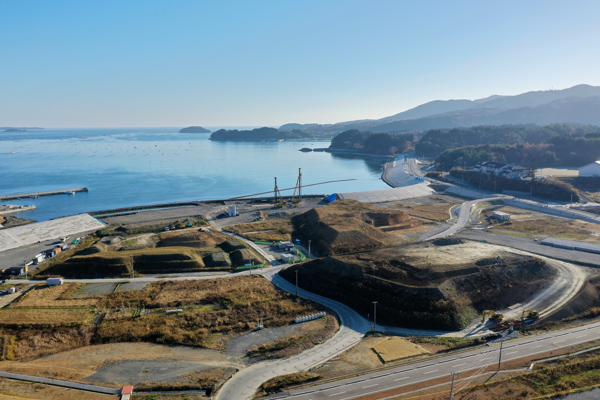 海岸沿いの風景で、手前には舗装された道路と建設中の土地が広がり、奥には穏やかな海と緑に覆われた山々が連なっている様子が見える写真