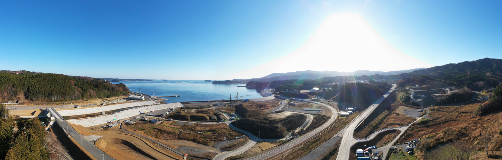 広い海とそれを囲む山々が背景に広がり、手前には工事現場や道路、車両が点在していて、自然と人の営みが混在する様子が見える写真