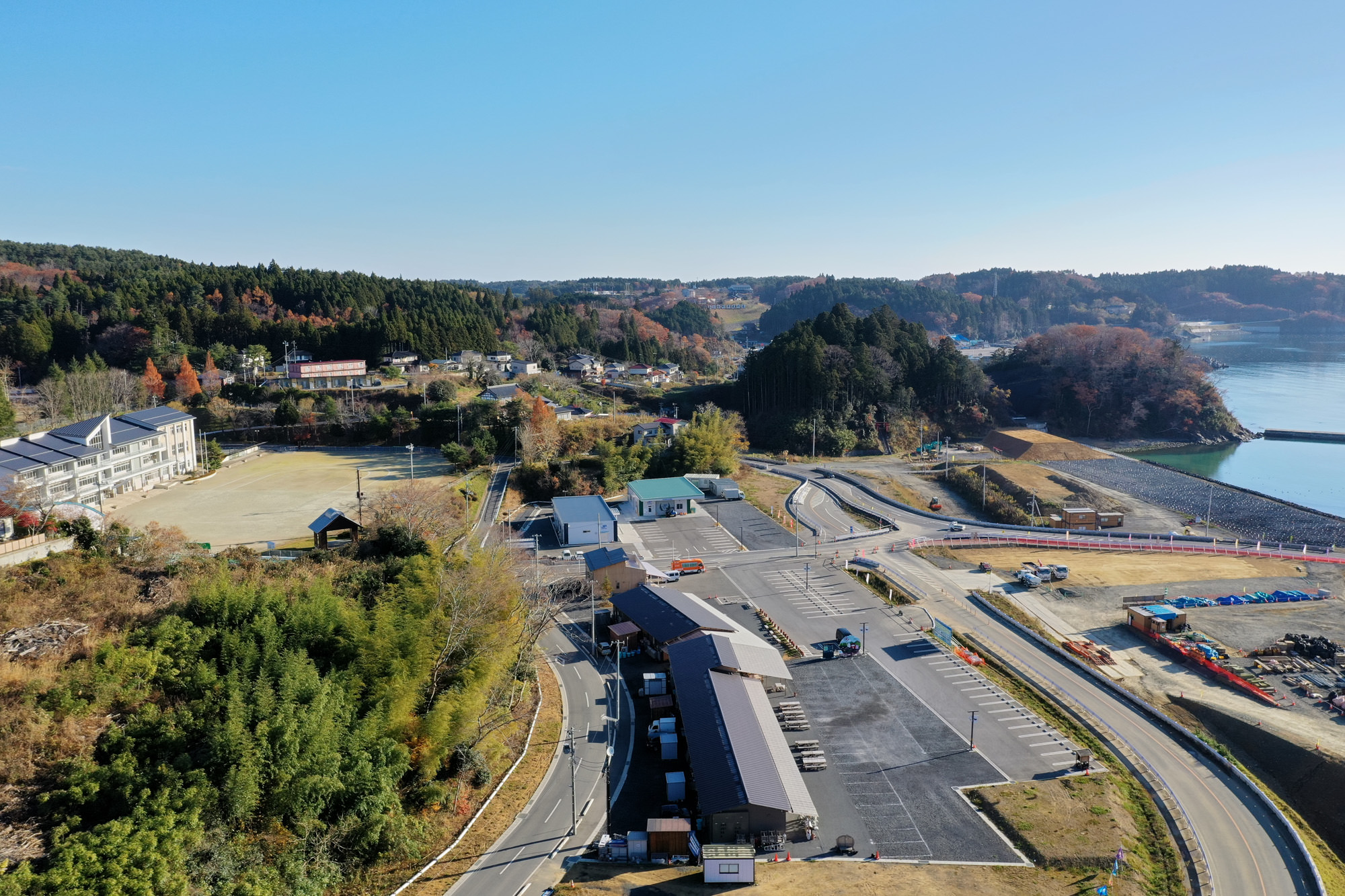 建物や駐車場、道路が広がる市街地が手前にあり、その周囲を緑豊かな木々が囲み、奥には森林に覆われた丘が連なり、右側には海岸線が広がって青い海と空が接している風景の写真