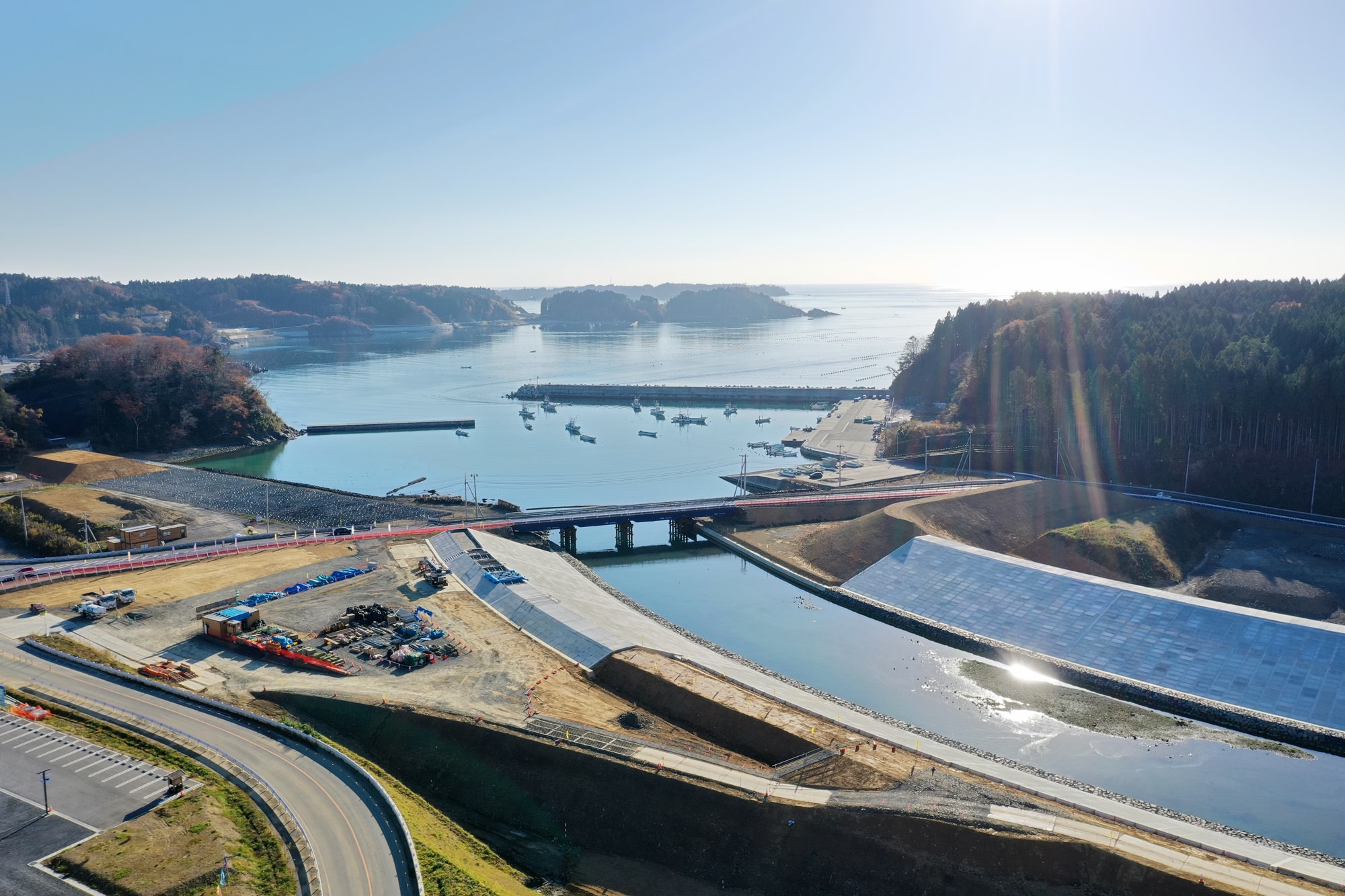 青い海にかかる橋の下に複数の船が浮かび、海岸線には木々と丘が広がり、晴れ渡った空の下で橋の近くには建設資材や機材が置かれている風景の写真