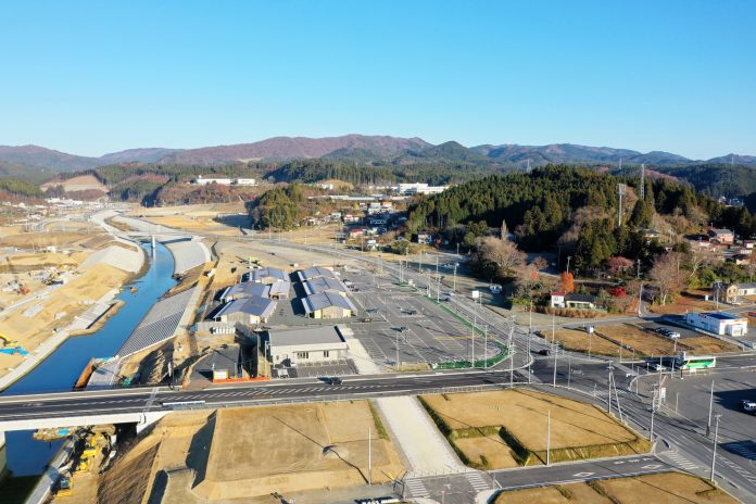 広々とした駐車場と複数の建物が並ぶ田舎の風景で、背景には緑に覆われた山々が連なっており、自然に囲まれた静かな地域の様子が見える写真