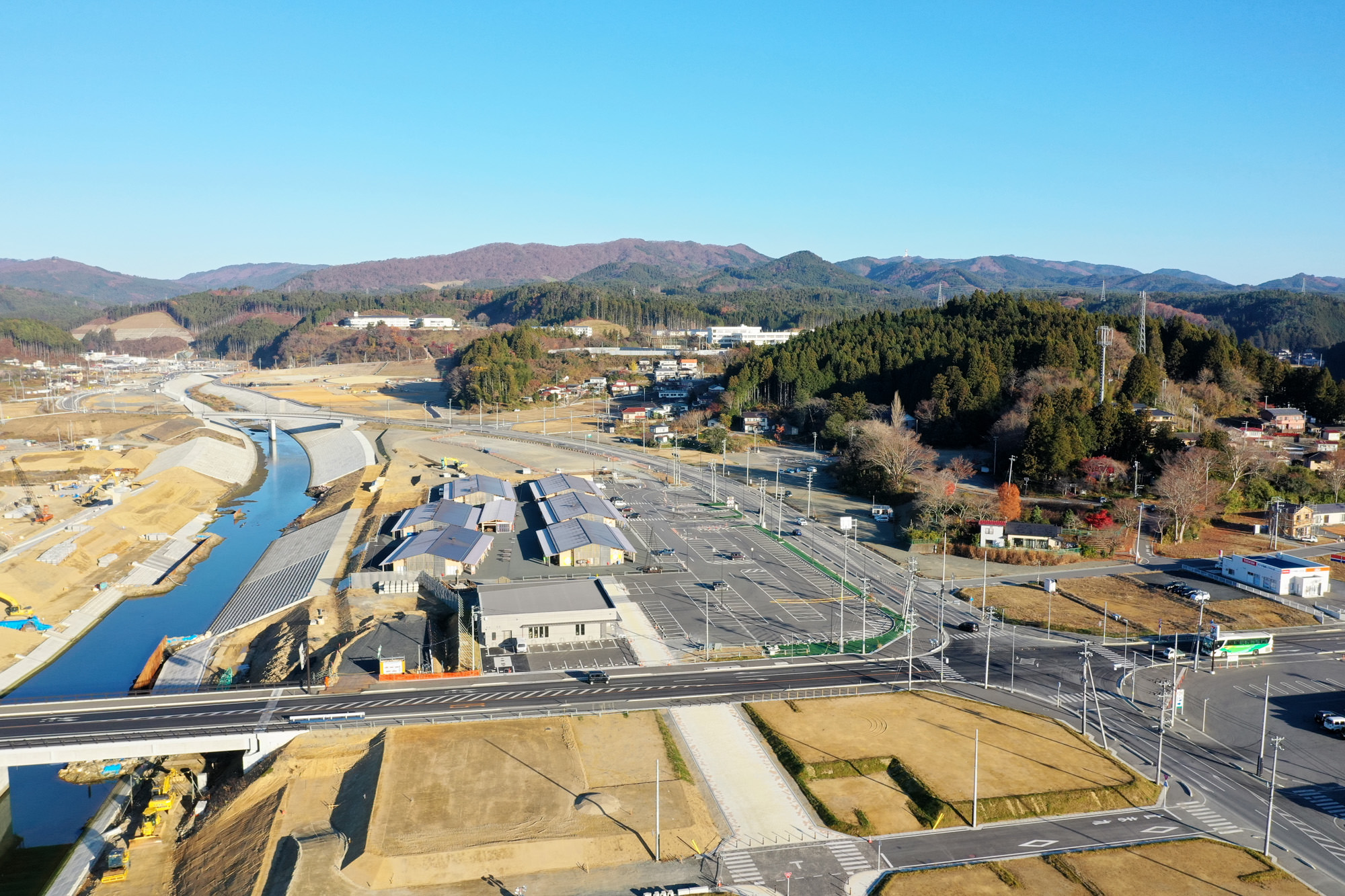 広々とした空と山々を背景に、川沿いに複数の建物と道路が整然と並び、自然と人の営みが調和している様子が見える写真