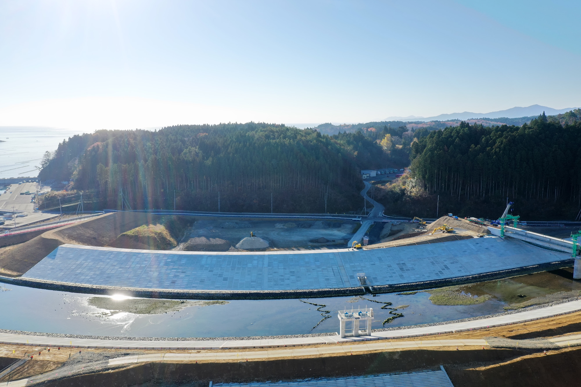 コンクリート製の大きなダムまたは堤防が水面の前にそびえ立ち、背景には木々に覆われた丘と青空が広がり、右側にはクレーンや足場などの建設設備が見える風景の写真