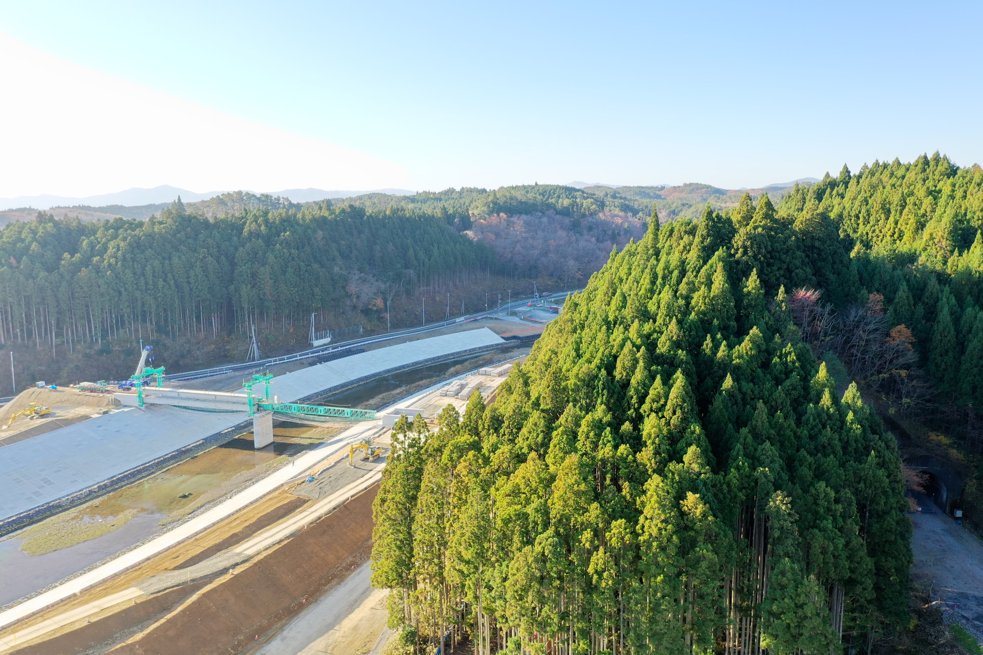 右側に背の高い常緑樹が密集する森林が広がり、左側には橋やコンクリート構造物を含む工事現場があり、背景には木々に覆われたなだらかな丘と青空が広がっている風景の写真