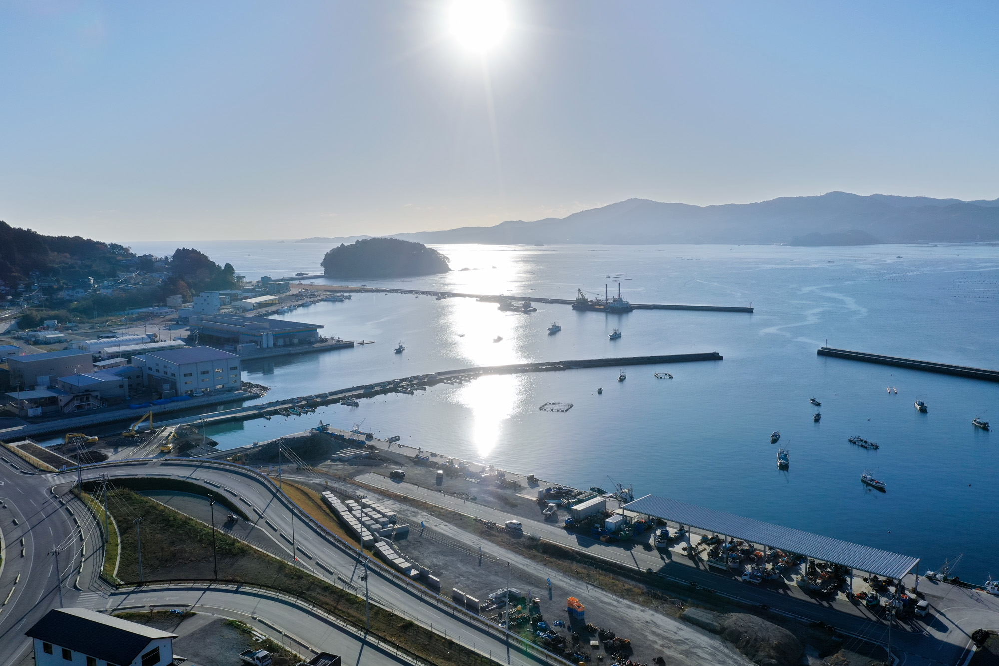 海岸沿いの町と港が広がり、穏やかな海面に太陽の光が反射してきらめき、自然の美しさと人の営みが調和している様子が見える写真