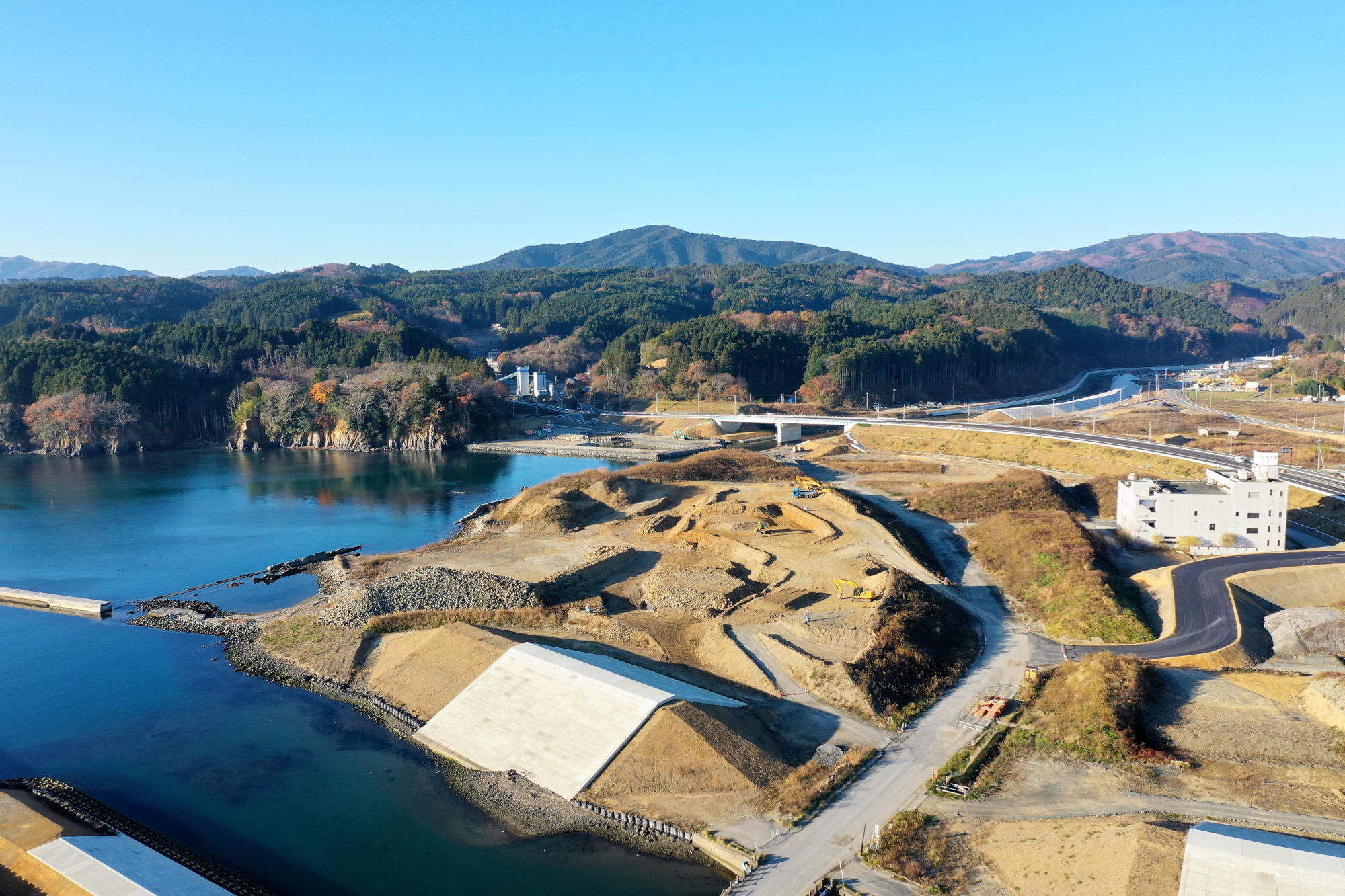 青い空の下、山々に囲まれた静かな湖のほとりに工事現場が広がっており、自然の中で開発が進められている様子が見える写真