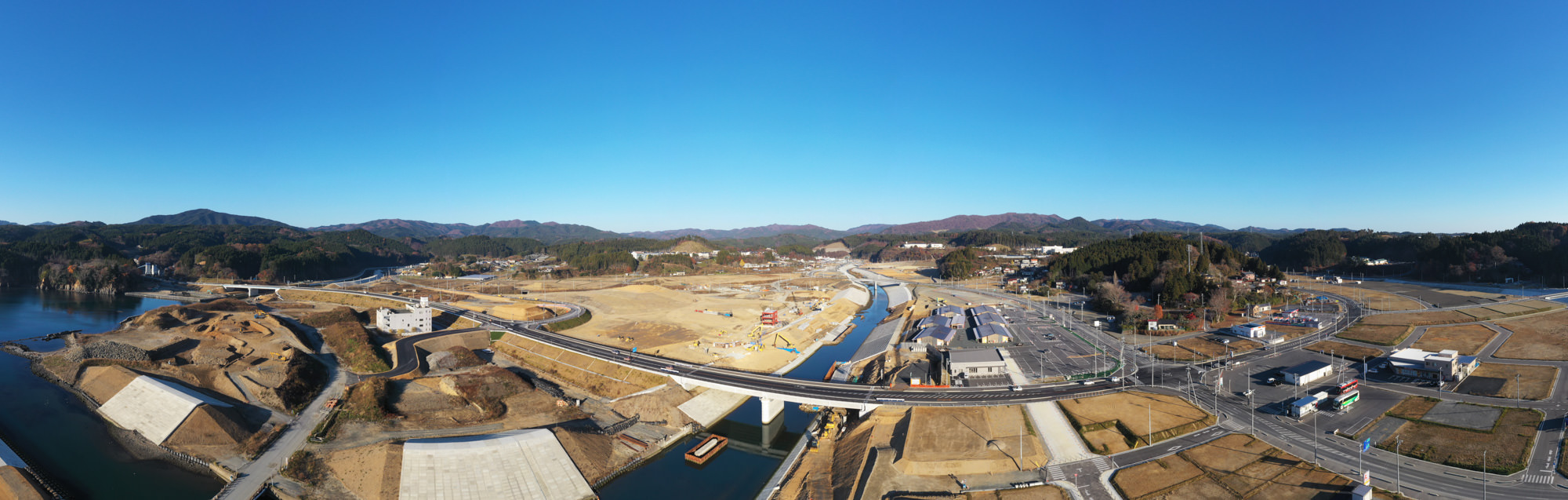 青空の下、山々に囲まれた広大な土地に川がゆったりと流れ、その上に橋が架かり、複数の道路が交差して自然と交通インフラが調和している様子が見える写真