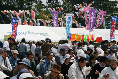 たくさんの鯉のぼりが空にたなびき、様々な幟やテントが立ち並ぶ賑やかな屋外のイベントで、多くの来場者たちで賑わっている様子の写真