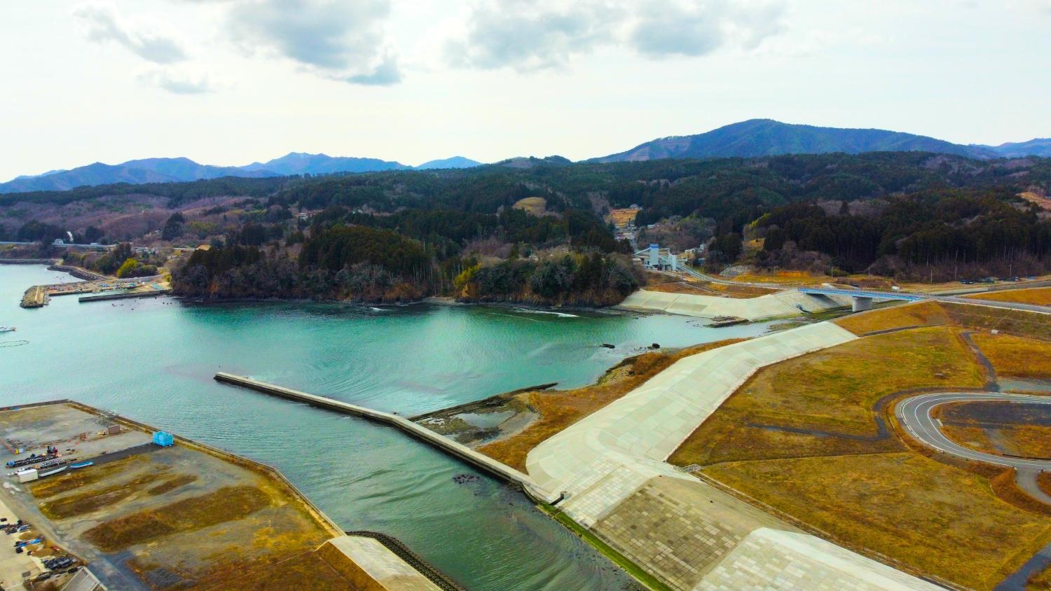 湾に注ぐ川の河口部が中央に位置し、左右には大きな防潮堤と整備された護岸が広がり、右手にはカーブした道路や造成された広い土地、奥には森林に覆われた山々と小さな集落が点在していて、自然と復興整備が共存する湾周辺の昼間の風景を高台から定点カメラで撮影した写真