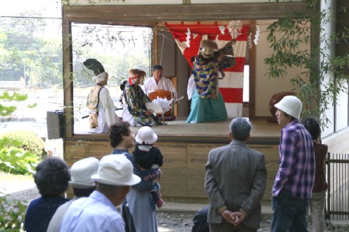 日差しの明るい屋外の舞台で、演者が華やかな衣装と面を着けて舞を披露し、数名の観客がその様子を熱心に鑑賞している様子の写真