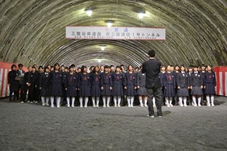 「祝 貫通 三陸沿岸道路 南三陸道路1号トンネル」と書かれた大きな横断幕、両側に紅白幕が掲げられたトンネル内で、制服姿の生徒たちが横に広がって並び合唱を行っている様子の写真