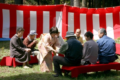 紅白幕が張られた屋外の会場に赤い毛氈が敷かれたベンチに観光客が座っており、着物姿の女性がお茶をもてなしている様子の写真