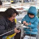 村山さんが指導しながらバーナーで熱し、屋外で水色のダウンジャケットを着た男の子が、長い金属棒の先についたドロドロになったオレンジ色のガラスの元の形を整えている、ガラス玉作成体験の様子の写真